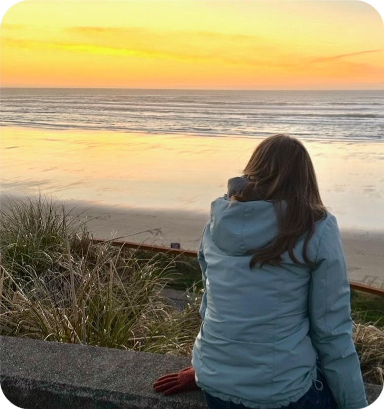 Kirsten Ellison sitting on a driftwood wall at a Pacific Northwest beach, watching a golden sunset over the ocean